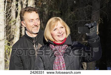 Stock Image - Mature couple taking a self-portrait with their camera in a forest; Canmore, Alberta, Canada. Fotosearch