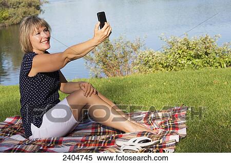 Stock Photography - Mature woman taking her picture with a cell phone while relaxing in Hawrelak Park; Edmonton, Alberta, Canada. Fotosearch