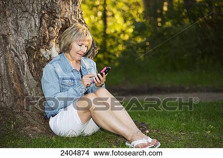 Picture - Mature woman using a cell phone in the park; Edmonton, Alberta, Canada. Fotosearch