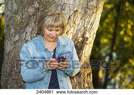 Stock Image - Mature woman using a cell phone in the park; Edmonton, Alberta, Canada. Fotosearch