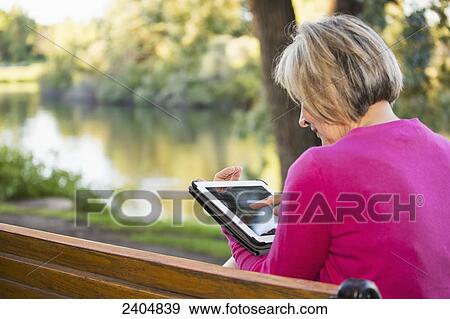 Stock Photo - Mature woman using a tablet in a city park; Edmonton, Alberta, Canada. Fotosearch