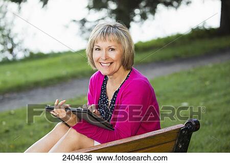 Stock Photograph - Mature woman using a tablet in a city park; Edmonton, Alberta, Canada. Fotosearch
