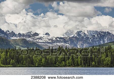 Stock Photo - Mt McKinley peaking through the clouds behind snowy rocky foothills of the Alaska range with Byers lake in the foreground, Denali State Park; Alaska, United States of America. Fotosearch