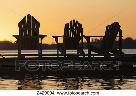 Picture - Silhouette of three adirondack chairs on a dock at sunset; Ontario, Canada. Fotosearch