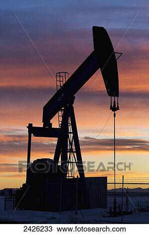 Stock Image - Silhouetted pumpjack at sunset with mountains in the background and glowing clouds; Alberta, Canada. Fotosearch