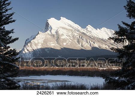 Snow covered mountain framed by silhouetted evergreen trees with snow covered lake and blue sky; Banff, Alberta, Canada View Large Photo Image Stock Image - Snow covered mountain framed by silhouetted evergreen trees with snow covered lake and blue sky; Banff, Alberta, Canada. Fotosearch