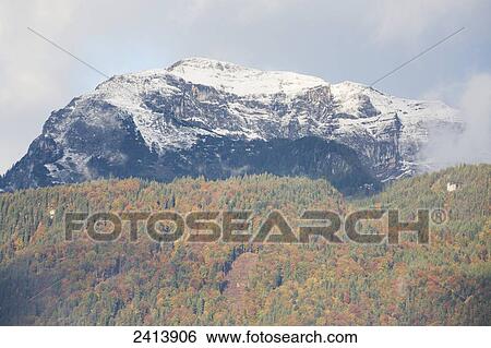 Stock Photograph - Snow covered rocky mountain peak viewed through cloud with blue sky and treed slope in autumn; Kramsach, Austria. Fotosearch