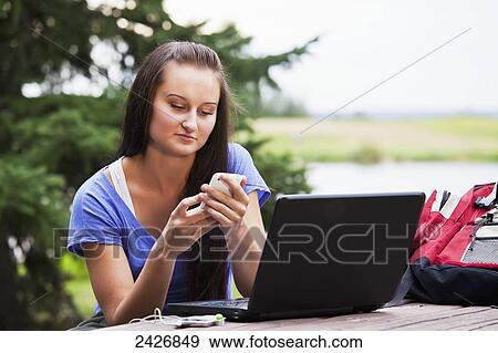 Stock Photo - Student checking for text messages while studying outdoors using a laptop; St. Albert, Alberta, Canada. Fotosearch