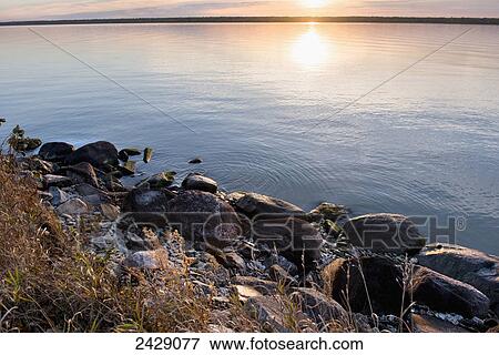 Stock Photo - The setting sun reflects on the tranquil water with rocks along the shoreline; Riverton, Manitoba, Canada. Fotosearch