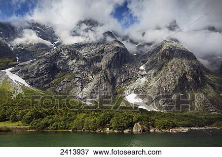 Stock Photo - Traces of snow on mountains along the water's edge with clouds covering the peaks; Andalsnes, Rauma, Norway. Fotosearch