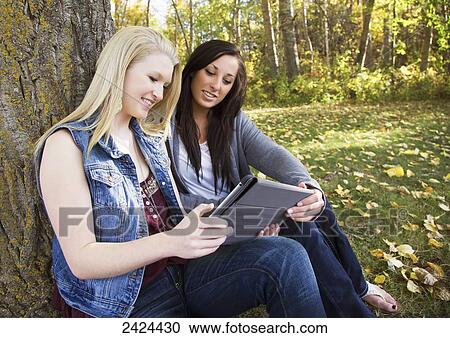 Two friends looking at a tablet in a park in autumn; Edmonton, Alberta, Canada View Large Photo Image Stock Image - Two friends looking at a tablet in a park in autumn; Edmonton, Alberta, Canada. Fotosearch