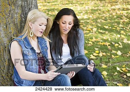Stock Photo - Two friends looking at a tablet in a park in autumn; Edmonton, Alberta, Canada. Fotosearch