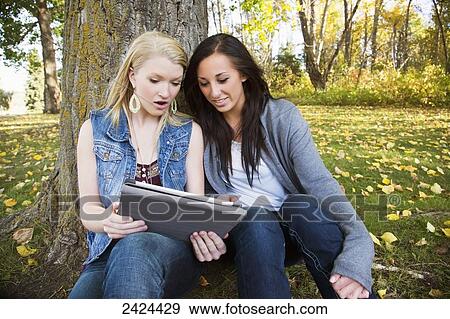 Stock Photo - Two friends looking at a tablet in a park in autumn; Edmonton, Alberta, Canada. Fotosearch