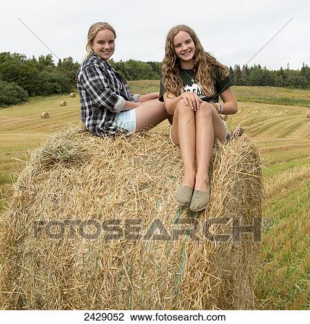 Stock Image - Two girls sit on top of a large hay bale in a field; Kensington, Prince Edward Island, Canada. Fotosearch