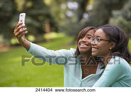 Two sisters taking a self-portrait with a smart phone in a park; Edmonton, Alberta, Canada View Large Photo Image Picture - Two sisters taking a self-portrait with a smart phone in a park; Edmonton, Alberta, Canada. Fotosearch