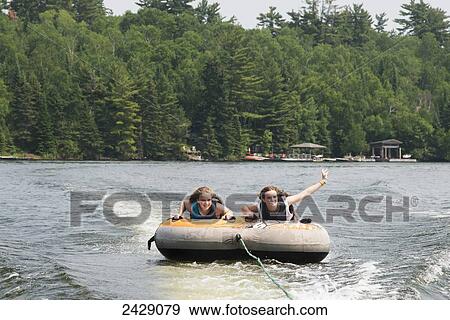 Stock Photo - Two teenage girls riding an inner tube being pulled behind a boat in a lake; Ontario, Canada. Fotosearch
