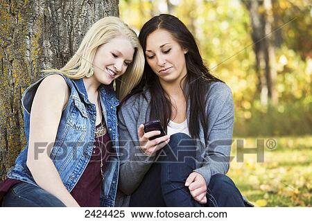 Stock Photography - Two teenage girls using a smart phone in a park; Edmonton, Alberta, Canada. Fotosearch