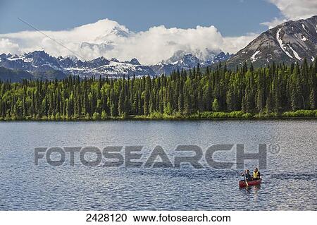 Stock Image - Two women and young girl in a red canoe on Byers lake with green forested shoreline and Mount McKinley peaking through the clouds, Denali State Park; Alaska, United States of America. Fotosearch