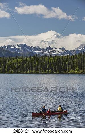 Stock Image - Two women and young girl in a red canoe on Byers lake with green forested shoreline and Mount McKinley peaking through the clouds, Denali State Park; Alaska, United States of America. Fotosearch