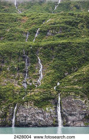 Picture - Waterfalls spilling down a green forested mountain side over a cliff into Passage cannal, Prince William Sound; Whittier, Alaska, United States of America. Fotosearch