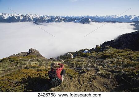 Stock Photo - Woman above fog bank photographing mountain goat (Oreamnos americanus) with Olympic Mountains in the background; Washington, United States of America. Fotosearch