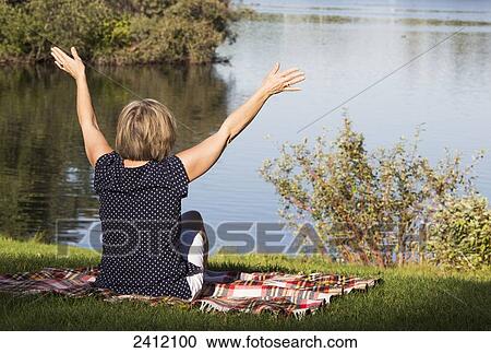 Stock Image - Woman sitting on a blanket in the park by a lake with arms raised in worship; Edmonton, Alberta, Canada. Fotosearch