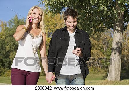 Stock Image - Young couple walking with cell phones in the park; Edmonton, Alberta, Canada. Fotosearch