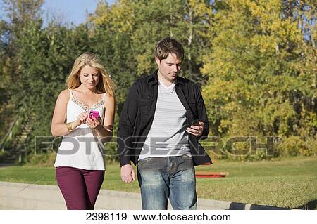 Young couple walking with cell phones in the park; Edmonton, Alberta, Canada View Large Photo Image Stock Photo - Young couple walking with cell phones in the park; Edmonton, Alberta, Canada. Fotosearch