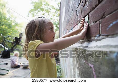 Stock Photo - Young girl rubbing coloured chalk into brick wall outside in her garden with her brother sitting behind her; Toronto, Ontario, Canada. Fotosearch
