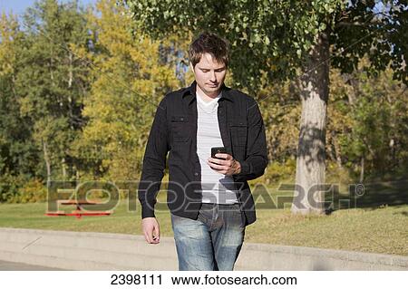 Stock Image - Young man with cell phone walking and texting; Edmonton, Alberta, Canada. Fotosearch