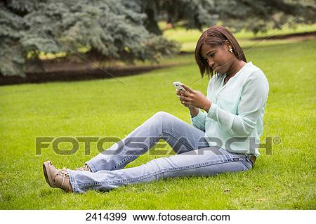 Stock Photo - Young woman on her smart phone in a park; Edmonton, Alberta, Canada. Fotosearch