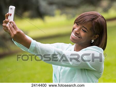 Stock Image - Young woman taking a self-portrait with her smart phone in the park; Edmonton, Alberta, Canada. Fotosearch