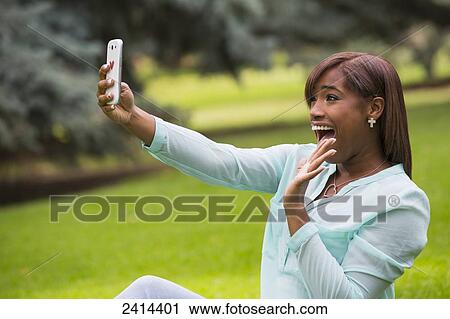 Stock Image - Young woman taking a self-portrait with her smart phone in the park; Edmonton, Alberta, Canada. Fotosearch