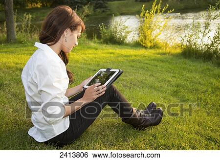 Stock Photograph - Young woman using her tablet in a park; Edmonton, Alberta, Canada. Fotosearch