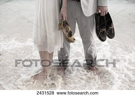 Stock Photo - A couple stand barefoot in the water holding shoes wearing formal clothing; Bridgetown, Barbados. Fotosearch