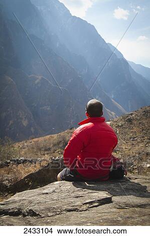 A man sits on a rock ledge overlooking the mountains of Leaping Tiger Gorge; Lijiang, Yunnan Province, China View Large Photo Image Picture - A man sits on a rock ledge overlooking the mountains of Leaping Tiger Gorge; Lijiang, Yunnan Province, China. Fotosearch