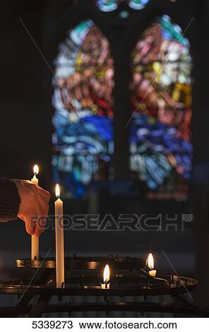 Stock Image - A man's hand lights a candle in a church with colourful stained glass window in the background; Northumberland, England. Fotosearch