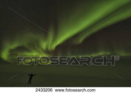 Stock Photograph - A person stands under the Northern Lights or Aurora Borealis; Old Crow, Yukon, Canada. Fotosearch