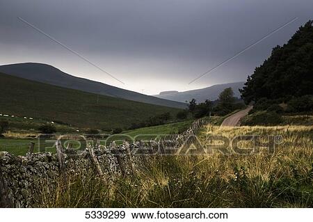 Stock Photo - A stone fence with wooden posts running along a road and the sun rising over the hills; Northumberland, England. Fotosearch