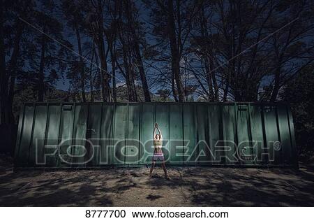 Stock Image - A woman doing a yoga pose under a light with a silhouette of trees and metal shipping container in the background; Tarifa, Cadiz, Andalusia, Spain. Fotosearch
