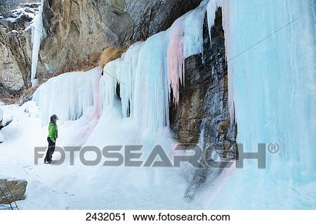 Stock Image - A woman hiker looks up at a frozen ice fall of pink and blue ice in Plateau Canyon; Palisade, Colorado, United States of America. Fotosearch