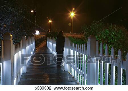 A woman stands on a wet boardwalk along an illuminated white picket fence; Sunderland, Tyne and Wear, England View Large Photo Image Stock Image - A woman stands on a wet boardwalk along an illuminated white picket fence; Sunderland, Tyne and Wear, England. Fotosearch