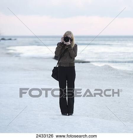 Stock Photograph - A woman stands on the beach with a camera pointed at the camera; Riverton, Manitoba, Canada. Fotosearch