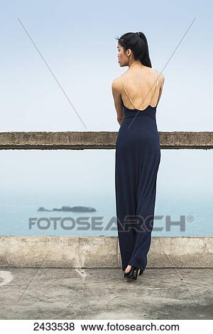 Stock Photo - A young woman in a formal dress stands at a railing looking out at the water; Xiamen, China. Fotosearch