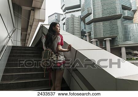 Stock Photo - A young woman stands on steps looking out at buildings; Hong Kong, China. Fotosearch