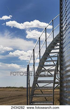 Stock Image - Close up of metal stair case on metal grain bin with clouds and blue sky; Alberta, Canada. Fotosearch