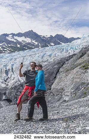 Stock Image - Couple taking a self-portrait with a cell phone in front of Shoup glacier, Shoup Bay State Marine Park, Prince William Sound; Valdez, Alaska, United States of America. Fotosearch