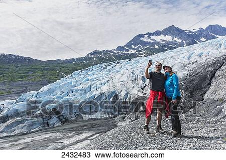 Stock Image - Couple taking a self-portrait with a cell phone in front of Shoup glacier, Shoup Bay State Marine Park, Prince William Sound; Valdez, Alaska, United States of America. Fotosearch