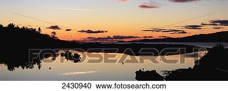Stock Image - Golden sunrise on the horizon with a silhouette of the shoreline reflected in a tranquil lake, Rosdohan Pier; Sneem, County Kerry, Ireland. Fotosearch