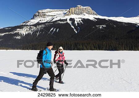 Stock Photography - Male and female snowshoeing on snow covered mountain lake with snow covered mountains and blue sky; Field, British Columbia, Canada. Fotosearch